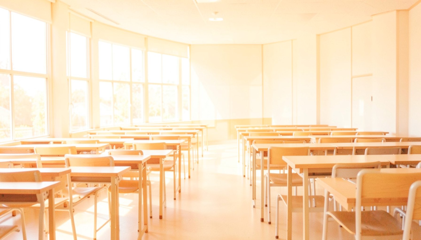 Aula luminosa con pupitres vacíos y grandes ventanales por donde entra luz natural, en tonos cálidos y suaves